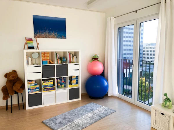 View in therapy room with a shelf in which are many games and therapy materials. Parquet floor, floor-to-ceiling window with a view of a building and trees.