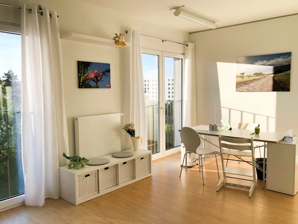 White curtains, two floor-to-ceiling windows, desk, chairs and two pictures on the white wall.